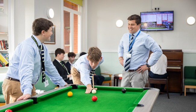 Three men in shirts and ties play snooker at an independent school in Sussex, whilst others sit and talk in the background; a piano and a television screen are also visible in the room.