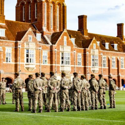 A group of people in camouflage military uniforms stand in formation on a grassy field outside a large, historic brick building—an independent school Sussex is proud to call its own, with many windows and tall chimneys.