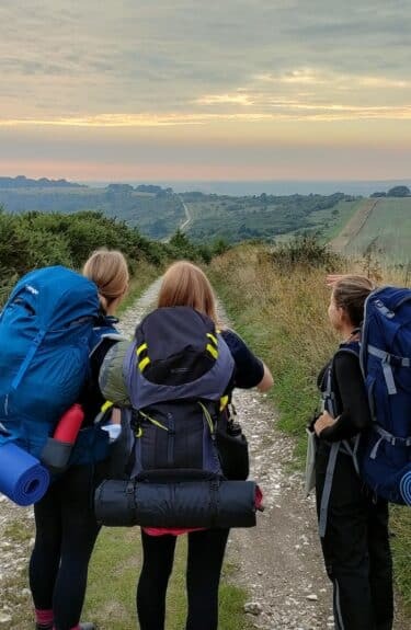 Three people with large rucksacks and camping gear stand on a gravel path, gazing at a sunset over green fields and hills near an independent school in Sussex. The partly cloudy sky creates a serene outdoor atmosphere.