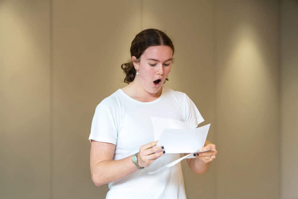A person wearing a white T-shirt holds and reads from sheets of paper with an expressive, open-mouthed look, possibly singing or acting, standing in front of a plain beige background—capturing the unique ethos of Eastbourne College.