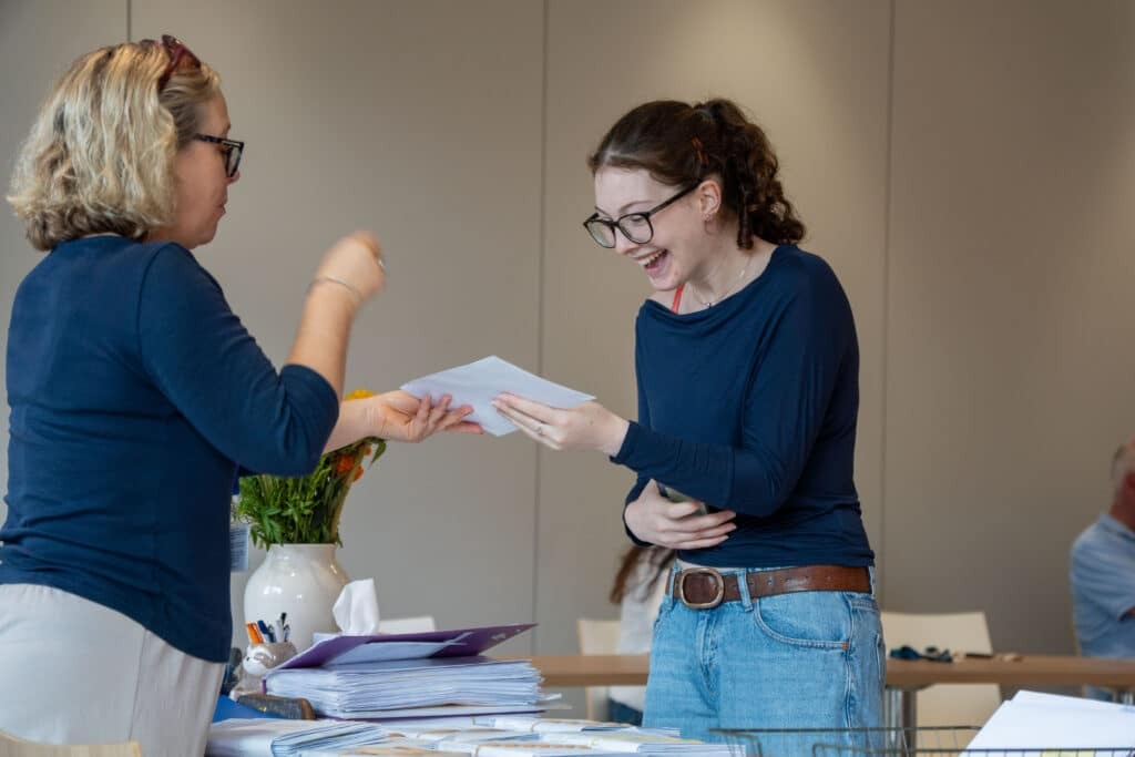 Two women stand by a table covered with papers, sharing A-level results. One hands a sheet to the other, who is smiling and happy. Both wear glasses and blue tops. There’s a vase of flowers on the table, reflecting Eastbourne College’s unique ethos.