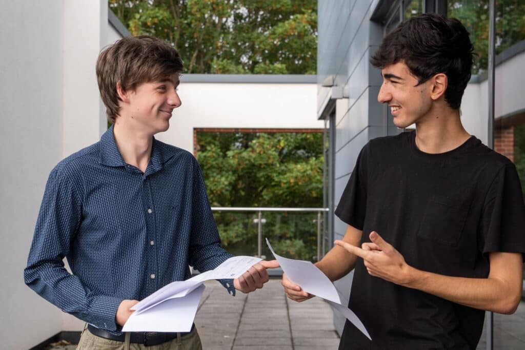 Two young men stand outside Eastbourne College, smiling and holding papers. Celebrating their A-level results, one points at the other's paper. Their joy captures the unique ethos of achievement amongst trees in the background.