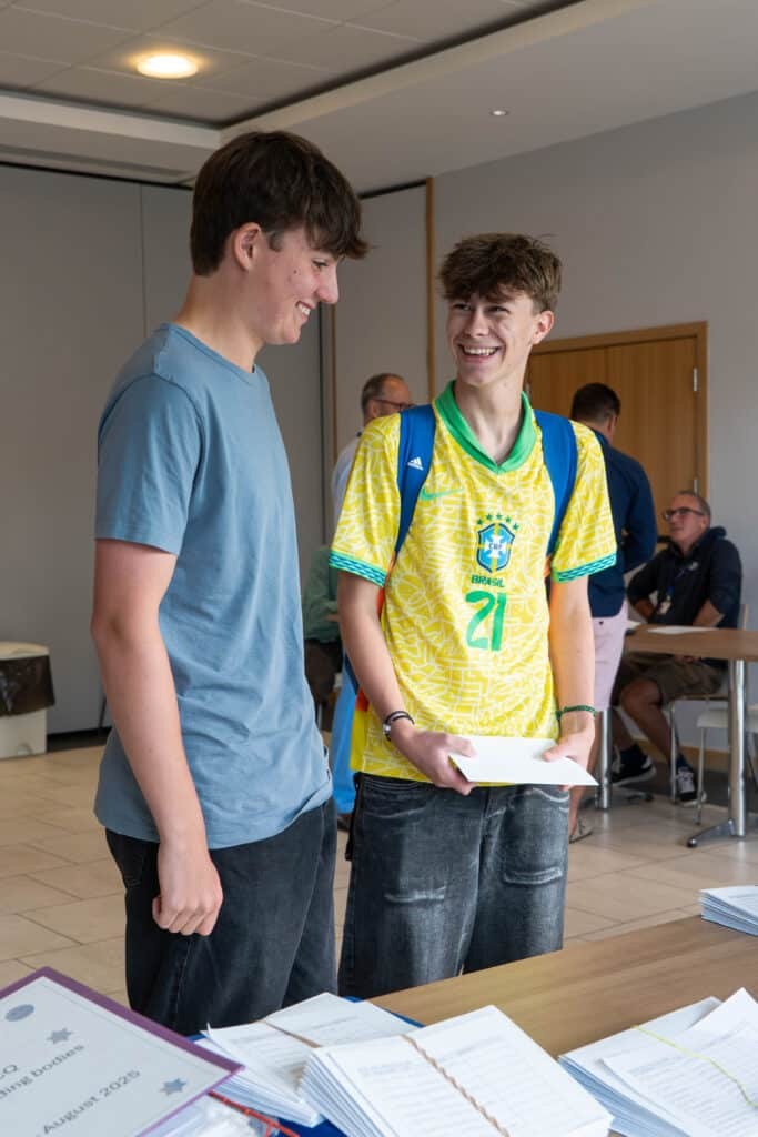 Two teenage boys stand indoors, smiling at each other. One wears a blue T-shirt and the other a yellow sports jersey, holding papers—likely GCSE results—at an Eastbourne College event, reflecting their ambitious approach to success.