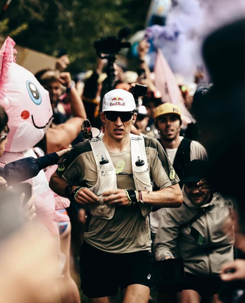 A male runner in a Red Bull cap and vest, resembling Tom Evans after his UTMB Victory, competes in a race surrounded by cheering crowds, photographers, and someone in a pink unicorn costume. The atmosphere is energetic and festive.