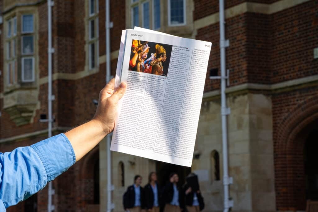 A person in a blue shirt holds up an open magazine, possibly the Tatler Schools Guide 2026, showing a page with text and a small colourful photo, standing outside a brick building with sporting successes featured in the background.
