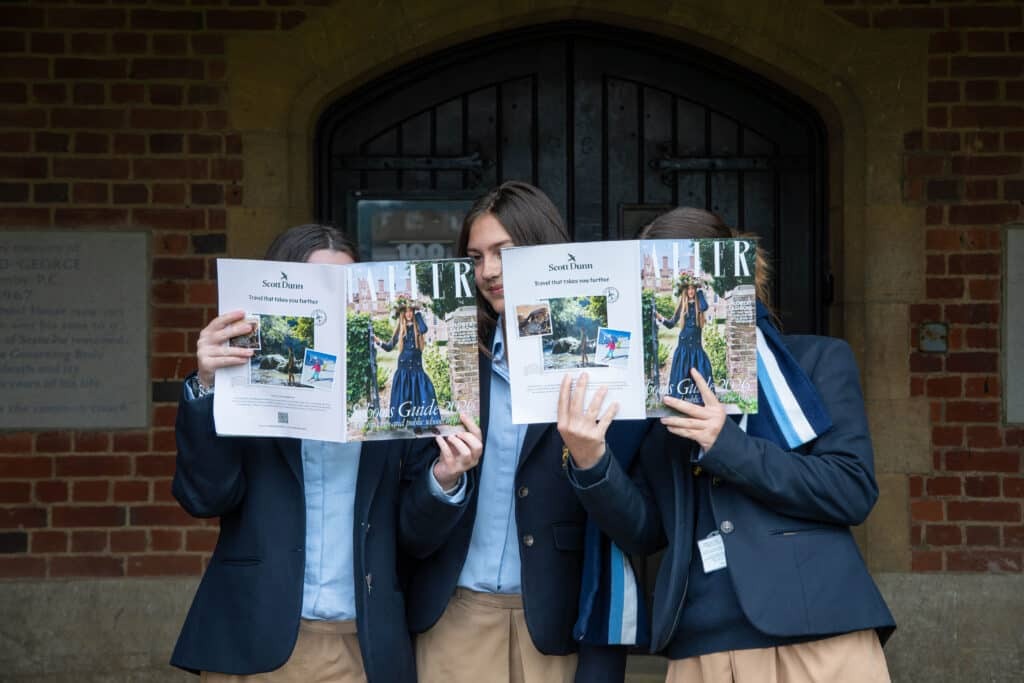 Three people in school uniforms stand side by side, holding magazines—including the Tatler Schools Guide 2026—in front of their faces, obscuring them. They are in front of a brick building with a dark wooden door, perhaps celebrating recent sporting successes.