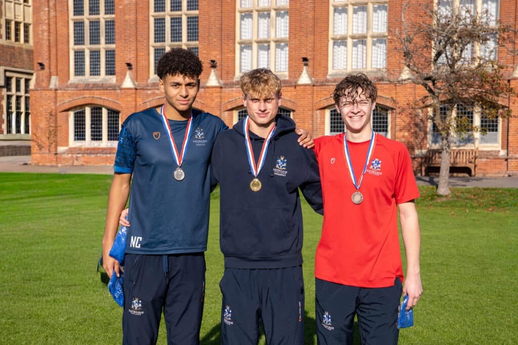 Three young men wearing medals stand arm-in-arm and smiling on a sunny lawn in front of a large brick building. Dressed in athletic trousers and Biathlon 2025 uniforms, they celebrate their achievements together.