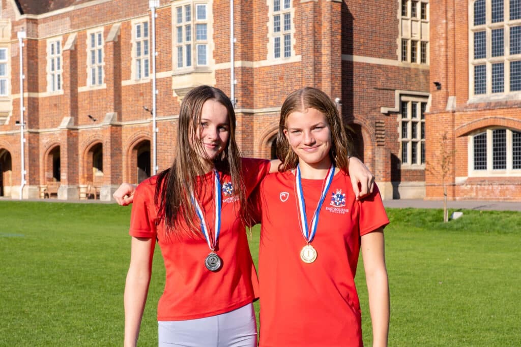 Two teenage girls with Biathlon 2025 medals around their necks stand side by side on grass in front of a brick school building, smiling at the camera and wearing red sports shirts.