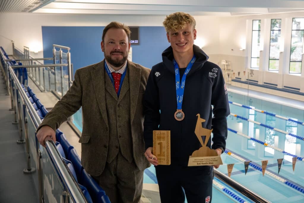 Two people stand by an indoor swimming pool; one is Lucas in a suit and medal, the other wears sportswear, a medal, and holds two wooden awards. Both are smiling at the camera after the Youth Euro Triathlon Championships.