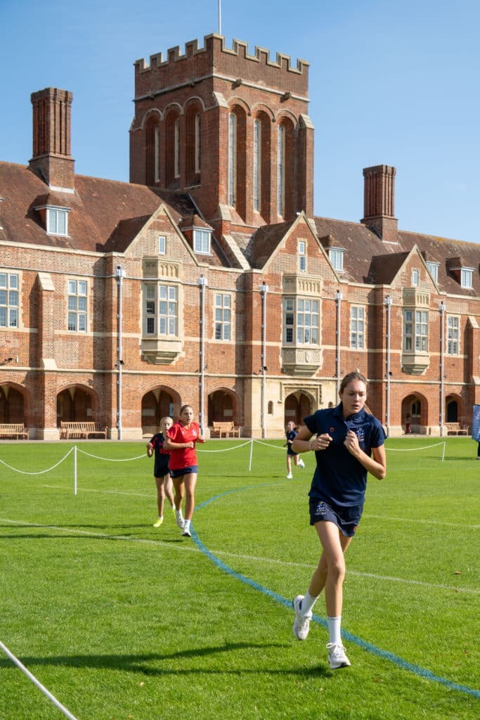 Three girls run on a grassy field in front of a large historic brick building with tall chimneys and arched windows, under a clear blue sky—capturing the energy of Biathlon 2025. The foreground runner wears navy, whilst the others wear red.