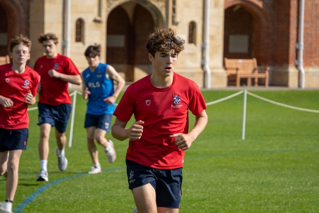 A group of teenage boys in sports kit are running on a grassy field near a historic brick building with arched doorways, training hard for Biathlon 2025. The boy at the front wears a red shirt and looks focused.