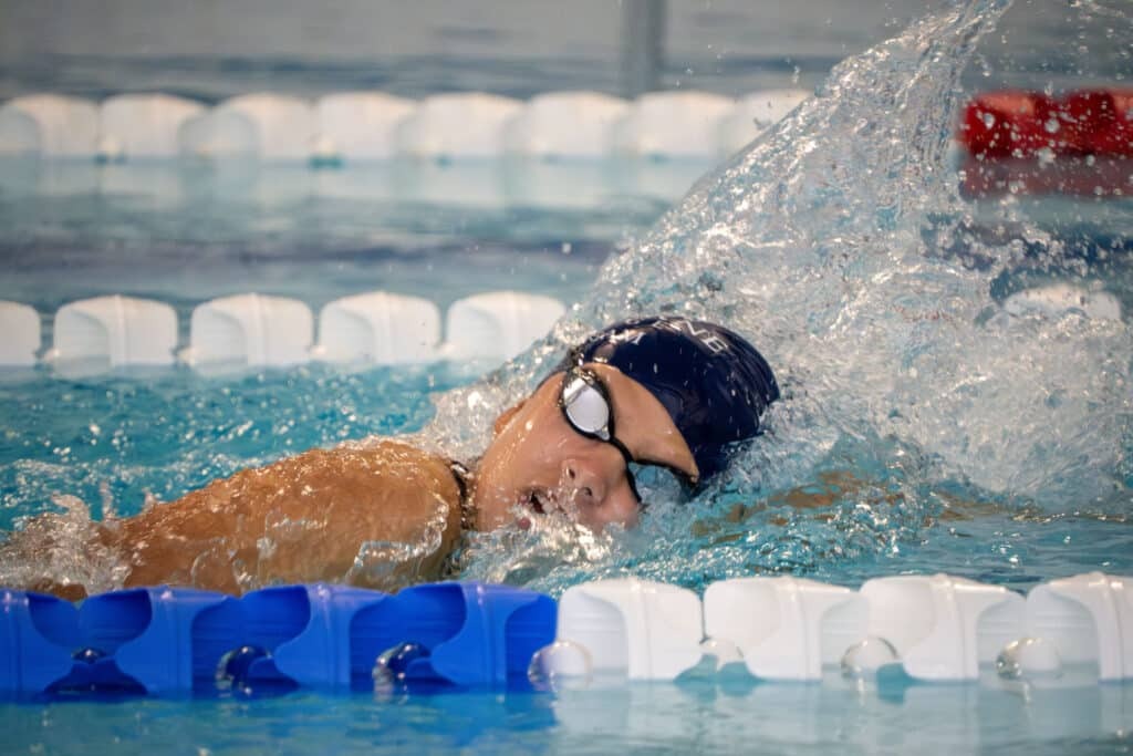A swimmer wearing goggles and a swim cap is doing front crawl in an indoor pool, turning their head to breathe with splashes of water around them and lane markers visible—training hard ahead of Biathlon 2025.
