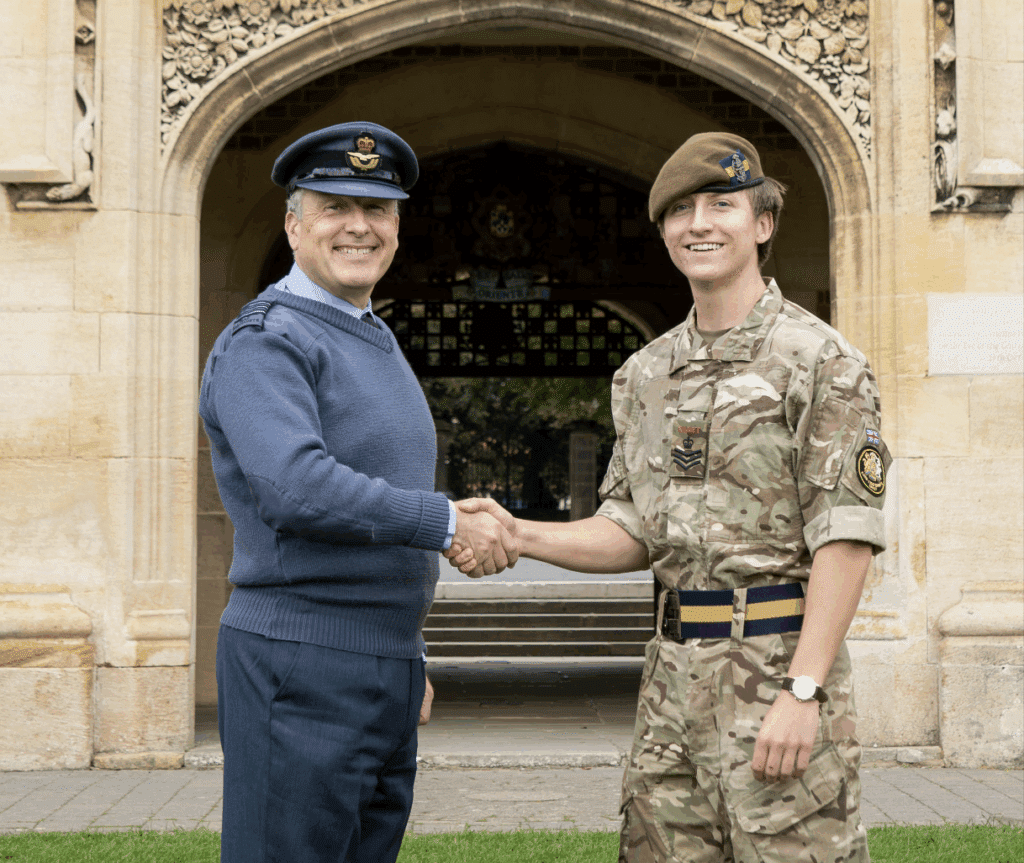 Two men in military uniforms shake hands and smile outdoors in front of an arched stone building. One, Henry, wears a blue Royal Air Force uniform; the other, a Lord Lieutenant’s Cadet from East Sussex, wears camouflage with a beret.