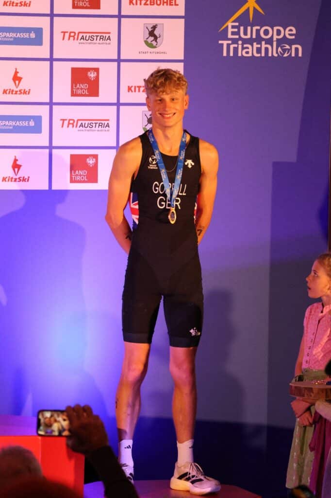 A male triathlete in a black GBR suit with a gold medal smiles on the podium at the European Triathlon Championships, as a young girl and someone taking a photo are seen in the foreground.