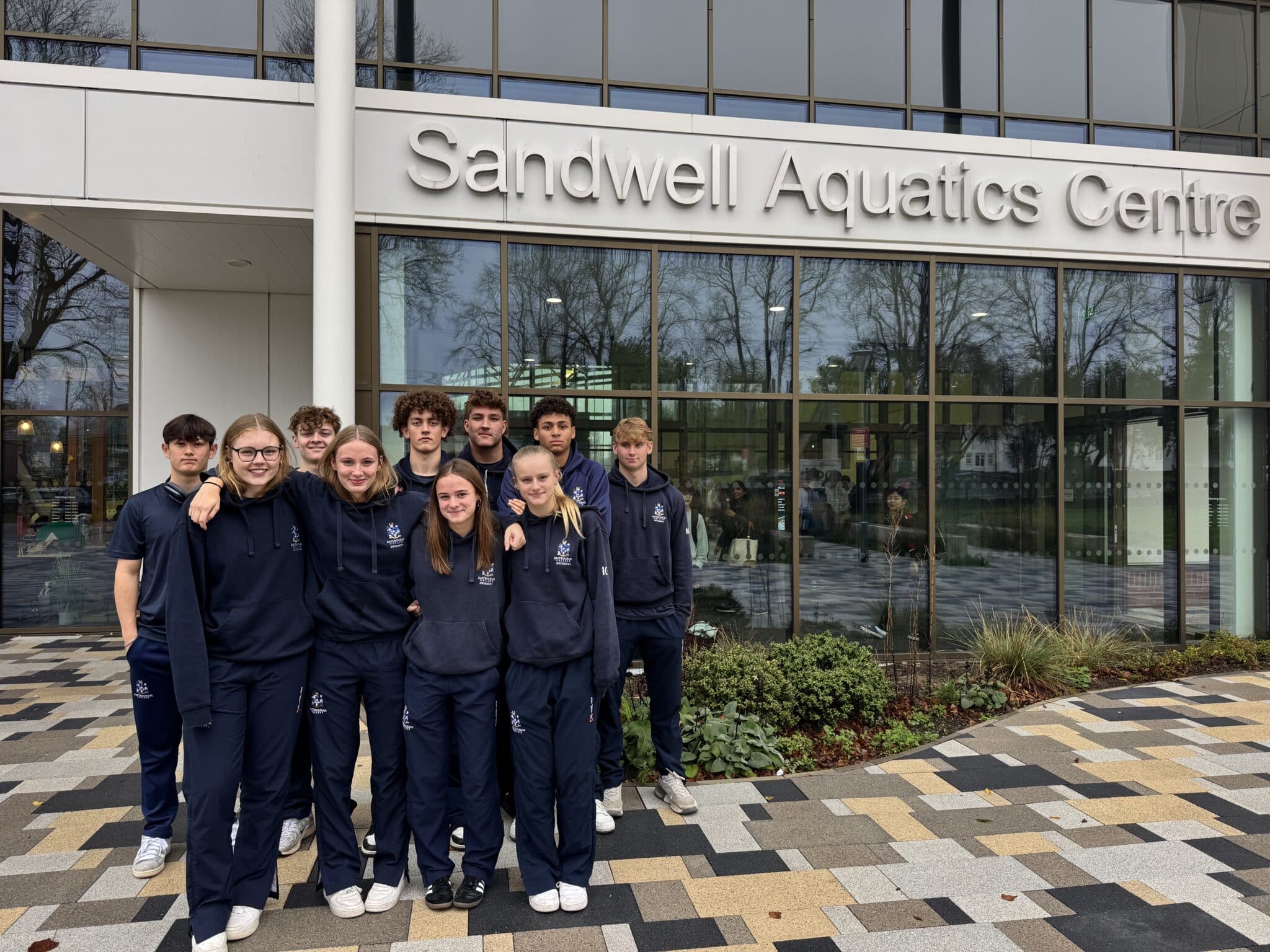 A group of young swimmers in matching navy tracksuits stands smiling outside the entrance of Sandwell Aquatics Centre, ready for the ESSA Secondary Team Championships, with large glass windows and the centre’s sign visible behind them.
