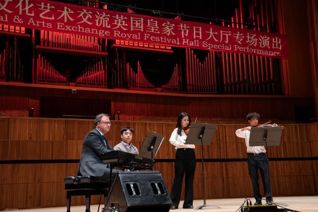 Four musicians perform onstage at Royal Festival Hall; one plays a keyboard whilst three others play wind instruments. A red banner above reads China–UK Youth Cultural Exchange Royal Festival Hall Special Performance in English and Chinese.
