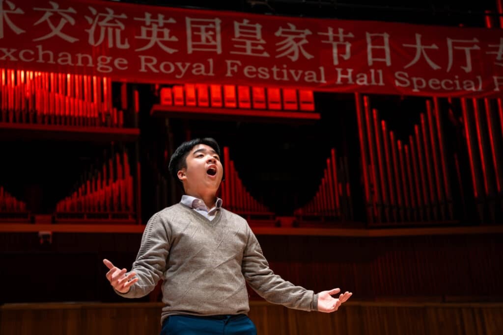 A man stands on stage with his arms open, singing passionately. Behind him are large organ pipes and a red banner with Chinese and English text reading China–UK Youth Cultural Exchange Royal Festival Hall Special.