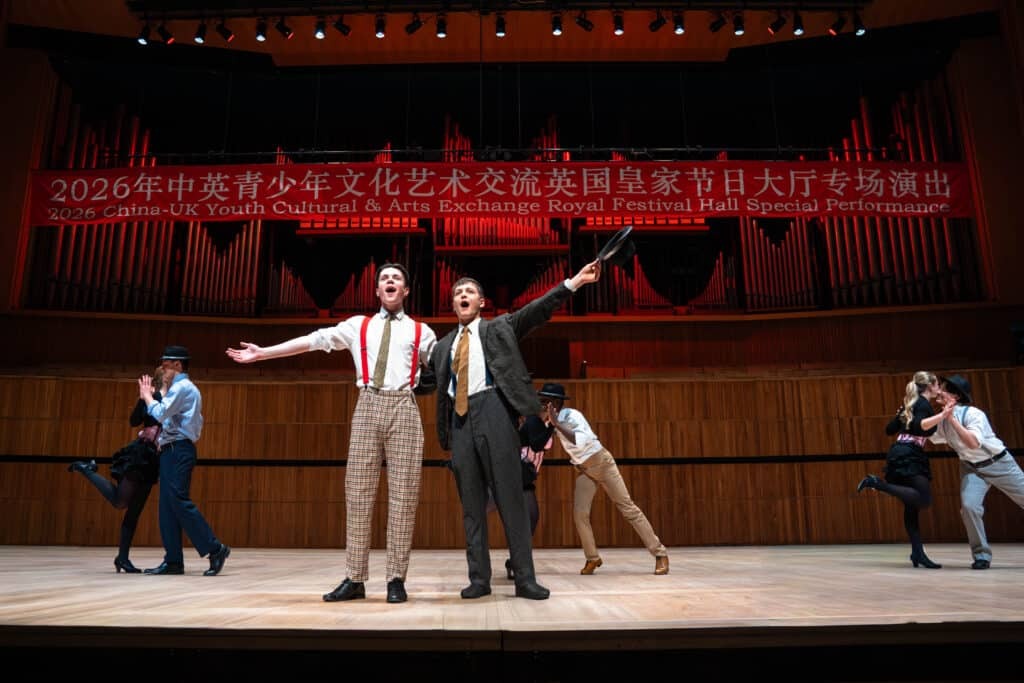 Two performers stand singing at centre stage, gesturing passionately, while pairs of dancers pose in the background. Above them is a red banner highlighting a China–UK Youth Cultural Exchange event at Royal Festival Hall and Eastbourne College.