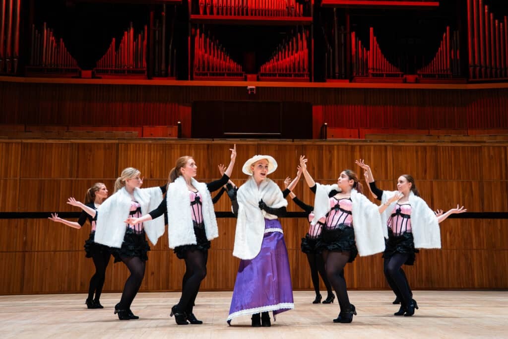 A group of women perform on stage at Royal Festival Hall; one stands centre in a purple skirt, white shawl, and hat, while five others in matching black-and-pink costumes and white shawls dance around her with raised arms.