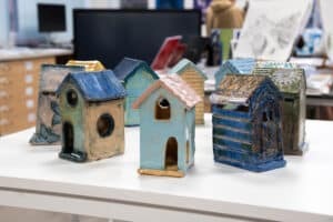 Several small, colourful ceramic houses with various shapes and roof styles are displayed on a white table in an art studio, reflecting the creativity celebrated by the Arts Society, with shelves and artwork visible in the background.