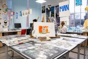 A bright art classroom with sketchbooks, drawings, and artworks displayed on tables and easels. Two students from the Visual Arts Society work at desks, surrounded by art materials and projects hanging on walls and lines.