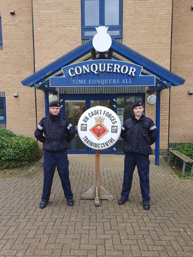 Two cadets in uniform stand on either side of an Army Cadet Force Training Centre sign, showcasing their training and discipline in front of a building with a blue sign reading CONQUEROR TIME CONQUERS ALL above the entrance.