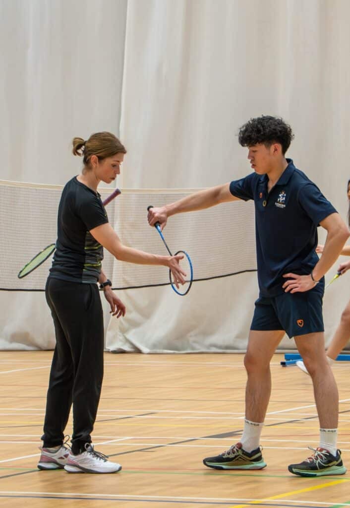 A woman, possibly inspired by Olympic Inspiration or even Heather Olver, coaches a young man on a badminton court. Both in sportswear, they stand near the net in an indoor gymnasium, perfecting his technique—perhaps at Eastbourne College.
