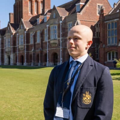 A person in a formal blazer with a crest stands on the grassy lawn of Eastbourne College, a historic brick building known for its arts and engineering excellence, featuring tall chimneys and arched windows under a clear blue sky.