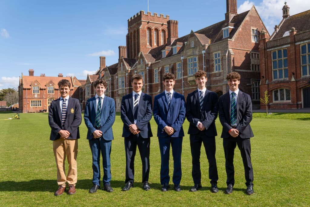 Six young men, Champions on the Pitch and proud Old Eastbournians, stand side by side on a lawn in suits and ties before a large historic brick school building beneath a blue sky with scattered clouds.