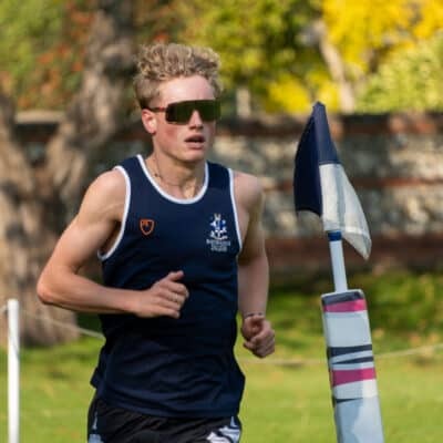 A young man in sports kit and sunglasses runs outdoors past a marker flag at the Eastbourne triathlon, with trees, grass, and a stone wall in the background.