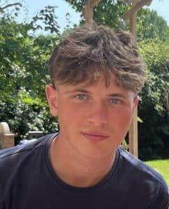 A young man with short, wavy brown hair and blue eyes is looking at the camera. Outdoors among green trees and sunlight, he wears a dark T-shirt, a chain necklace, and has an air of confidence befitting an Old Eastbournian like Freddy Powis.