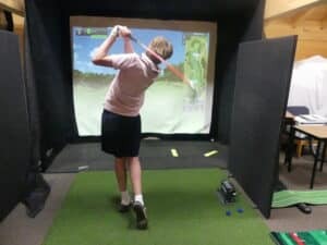 A person swings a golf club in an indoor golf simulator at Gonville House, aiming at a digital course projected on a screen. Green artificial turf and golf equipment are visible as part of the 24 Sports experience.