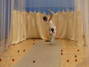 A cricket player from Gonville House in full protective kit bats in an indoor practice net. Multiple red cricket balls are scattered on the floor around the mat, and beige curtains hang behind the sports player.