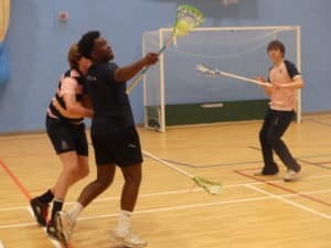 Three young people play indoor lacrosse at Gonville House; one player in dark shorts swings a stick while being defended by a player in pink and navy, as a third watches near the goal during this fast-paced sports match.