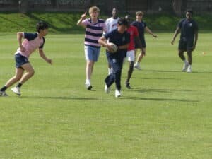 A group of boys in sports kits from Gonville House are playing rugby on a green field, enjoying the thrill of sport under the open sky.
