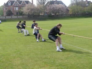A group of boys from Gonville House playing tug of war on a grassy field, pulling on a rope with effort. Houses and trees with blooming flowers are visible in the background during the 24 sports event.