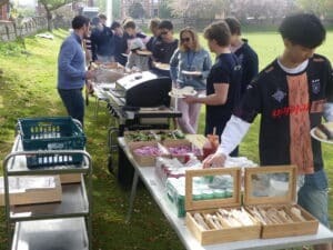 A group of people stand in a queue at an outdoor Gonville House barbecue, serving themselves food from tables with salads, cutlery, and condiments on a grassy area in daylight. Some are holding plates and chatting.