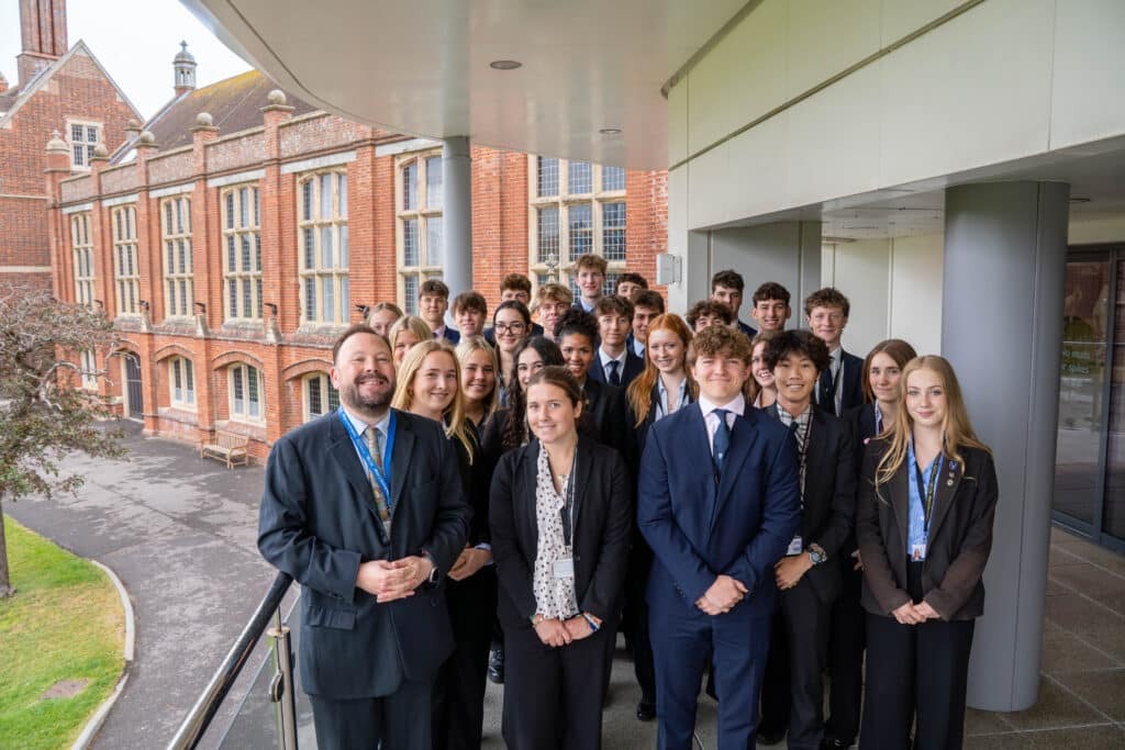 A group of students and two adults in business attire stand and smile on a balcony outside a brick school building. The group is posing for a formal photo on a cloudy day.
