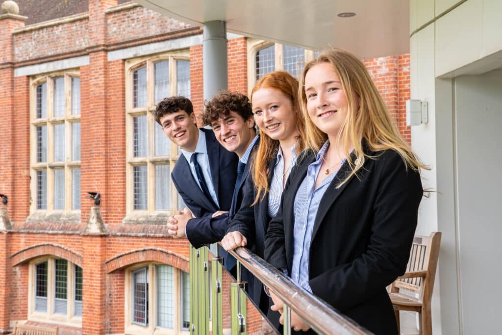 Four young adults wearing suits stand on a balcony, smiling at the camera. Behind them is a brick building with large windows and arched details, suggesting an academic or professional setting.