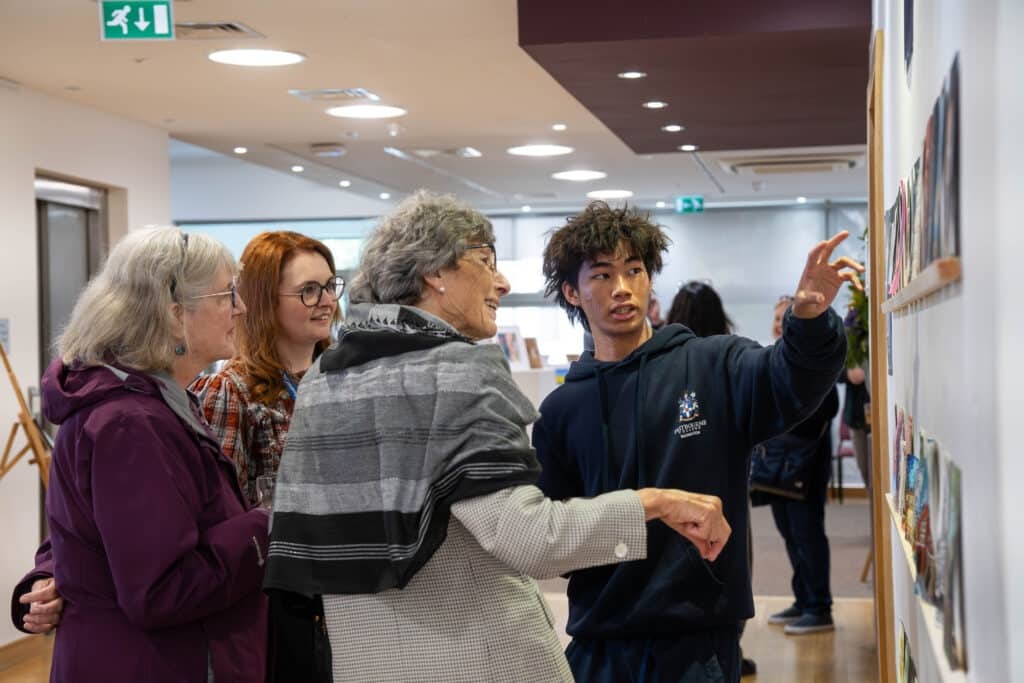 A young man points to artwork on a wall while three older women stand beside him, smiling and engaged. They appear to be having a friendly conversation in a bright, modern gallery space.
