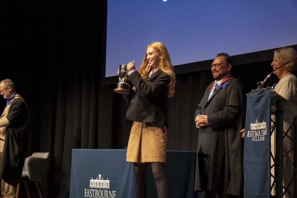 A young woman in a school uniform holds up a trophy on stage during Eastbourne College’s Speech Day, a true celebration of achievement, whilst three adults in academic robes stand nearby, smiling.