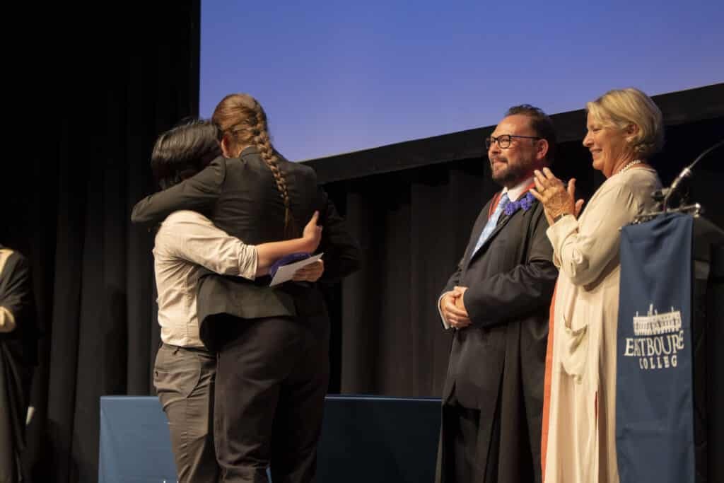 Two people embrace on stage during Speech Day at Eastbourne College, while two others stand nearby smiling and applauding. A lectern with the college logo is visible on the right, highlighting this moment of achievement.