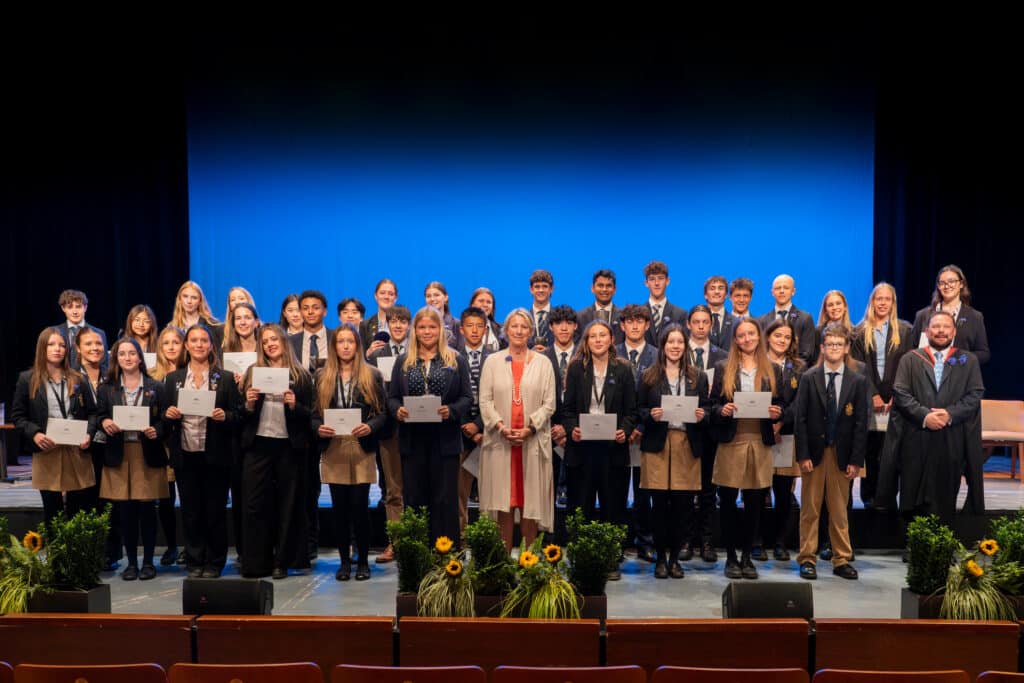 A large group of students and teachers stands onstage at Eastbourne College’s Speech Day, dressed formally. The students hold achievement certificates and smile at the camera, with plants and flowers arranged along the blue-lit stage.