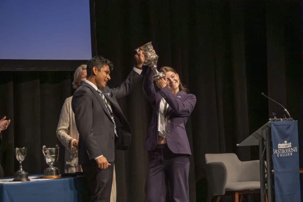Two people in suits stand on stage, raising a large trophy together and smiling—celebrating their achievement at Eastbourne College’s Speech Day. Other trophies sit on a nearby table, with black curtains hanging in the background.