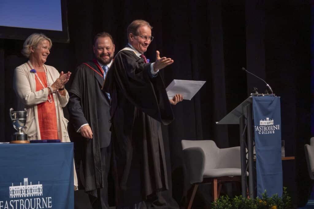 A man in academic robes gestures and smiles on stage at a graduation ceremony during Eastbourne College Speech Day, holding a certificate. Two colleagues beside him applaud as part of this Celebration of Achievement. The stage is decorated with banners.