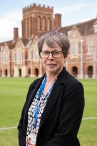 A woman with short grey hair and glasses, wearing a black blazer and patterned blouse, stands on a green lawn in front of a large historic brick building. She is wearing a blue lanyard with an ID badge.