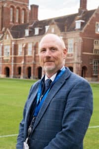 A middle-aged man with a light beard and short hair, wearing a blue checked suit, tie, and lanyard, stands on a lawn in front of a large, historic brick building with tall windows and chimneys.