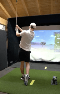 A golfer wearing a white shirt, black shorts, and a cap swings a club indoors on artificial turf, facing a golf simulator screen displaying virtual course graphics.