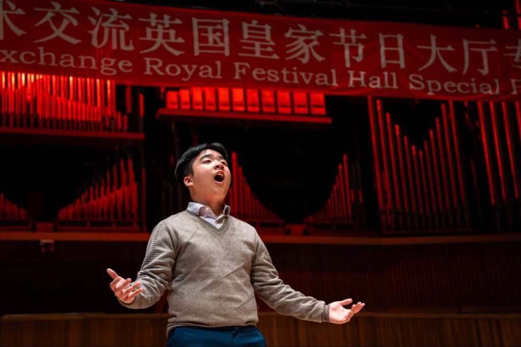 A man stands on stage with his mouth open, appearing to sing passionately. Behind him is a large red banner with Chinese and English text and organ pipes, suggesting a performance or festival at a concert hall.