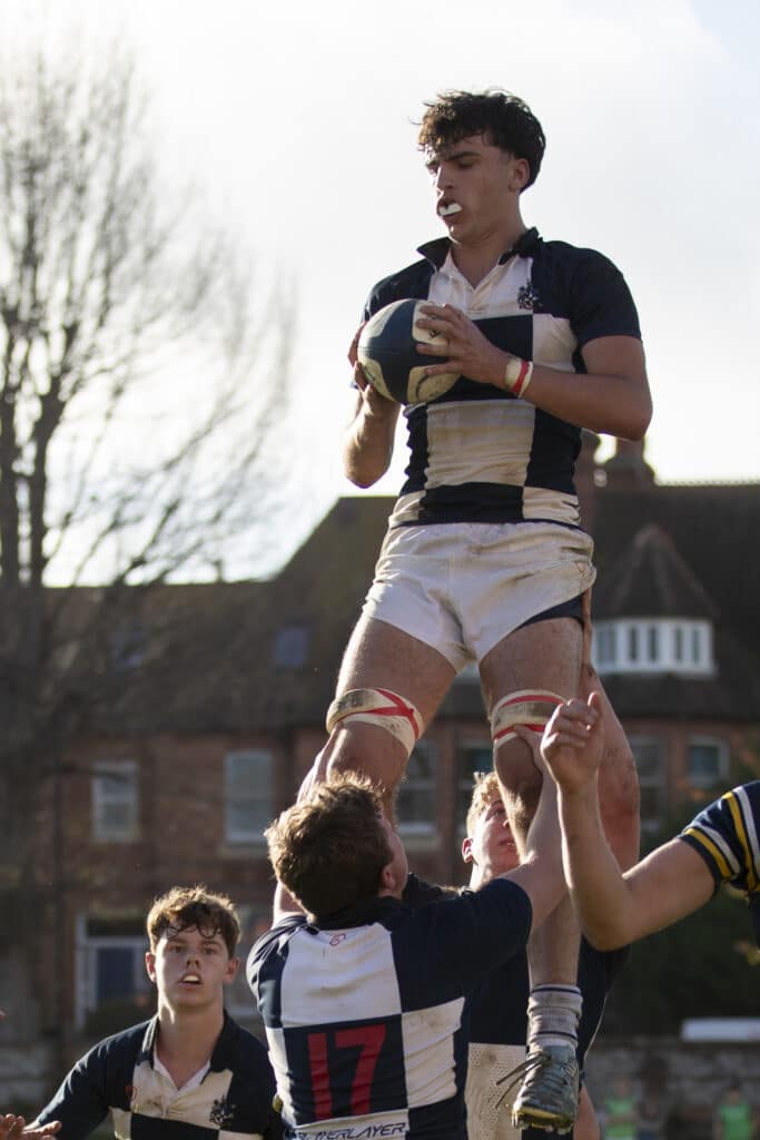 A rugby player is lifted by teammates to catch the ball during a line-out, with sunlight illuminating the fine art of athleticism and buildings visible in the background—a moment reminiscent of A-level success at Eastbourne College.